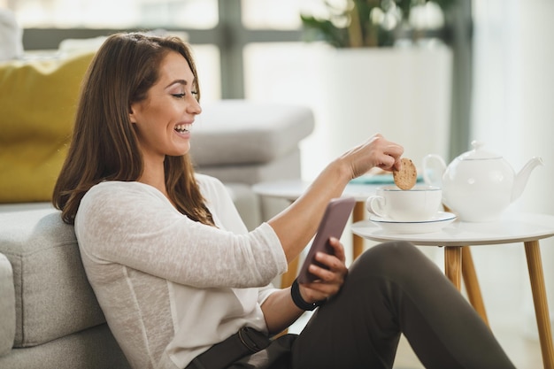Piękna, młoda kobieta cieszy się czasem na herbatę, trzymając smartfon i surfując po mediach społecznościowych w swoim domu.