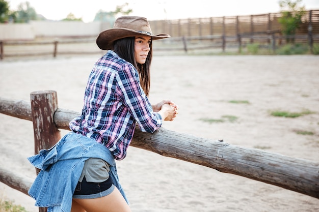 Piękna młoda kobieta cowgirl w kapeluszu i koszuli w kratę, stojąc i myśląc