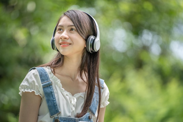 Piękna młoda kobieta dziewczyna w beżowej koszuli pozuje w zielonym mieście Park w tle Uśmiechnięta dziewczyna Słuchanie muzyki ze słuchawkami uczucie Dotykanie szczęścia Koncepcja stylu życia ludzi Makieta miejsca kopiowania