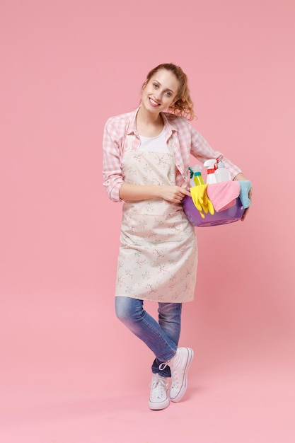 Piękna młoda kobieta gospodyni w fartuch trzymać umywalkę z butelek detergentu mycie środków czyszczących robienie prac domowych na białym tle na pastelowym różowym tle portret studio. Koncepcja sprzątania. Patrząc na aparat.