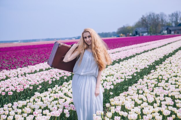 Piękna młoda kobieta jest ubranym w biel sukni pozyci z bagażem na kolorowym tulipanu polu z długim czerwonym włosy.