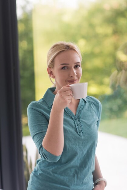 piękna młoda kobieta pijąca poranną kawę przy oknie w swoim domu