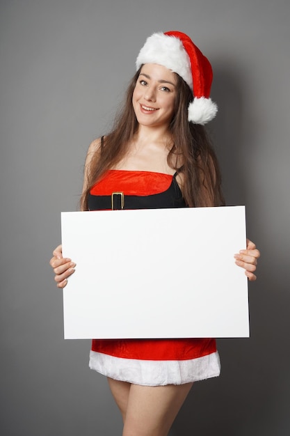 piękna młoda kobieta po dwudziestce ubrana w bożonarodzeniowy kostium, czerwoną sukienkę i kapelusz Świętego Mikołaja, przedstawiająca pusty znak z miejscem na kopię
