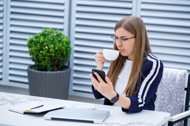 Piękna młoda kobieta pracuje na laptopie i rozmawia przez telefon, uśmiechając się siedząc na świeżym powietrzu w kawiarni. Młoda kobieta za pomocą laptopa do pracy. Freelancerka pracująca na laptopie w kawiarni na świeżym powietrzu