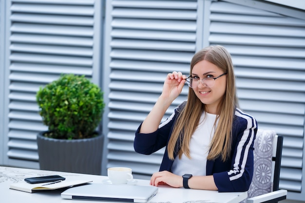 Piękna młoda kobieta pracuje na laptopie i uśmiecha się siedząc na świeżym powietrzu w kawiarni. Młoda kobieta za pomocą laptopa do pracy. Kobieta freelancer pracuje na laptopie w kawiarni na świeżym powietrzu.