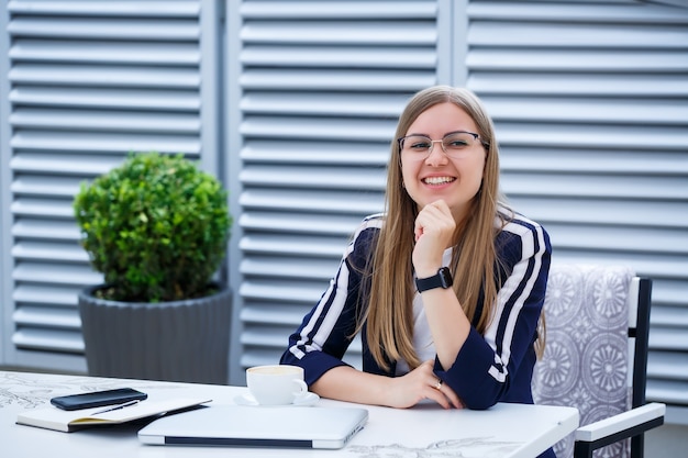 Piękna młoda kobieta pracuje na laptopie i uśmiecha się siedząc na świeżym powietrzu w kawiarni. Młoda kobieta za pomocą laptopa do pracy. Kobieta freelancer pracuje na laptopie w kawiarni na świeżym powietrzu.