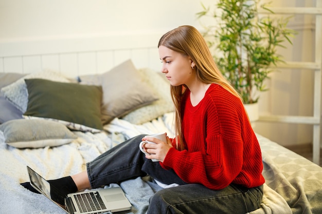 Piękna Młoda Kobieta Pracuje Na Laptopie, Siedząc Na łóżku W Domu, Pije Kawę I Uśmiecha Się. Praca W Domu Podczas Kwarantanny. Dziewczyna W Swetrze I Dżinsach W Domu Na łóżku