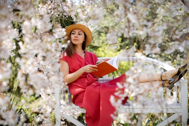 Piękna młoda kobieta siedzi na eleganckiej ławce w wiosennym ogrodzie pod kwiatem wiśni i czyta książkę