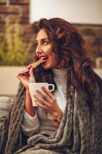 Piękna młoda kobieta siedzi na sofie wsuniętej w miękki koc, pije poranną kawę i gryzie ciasteczko.