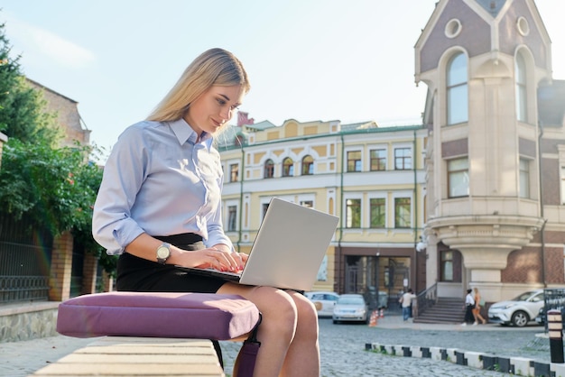 Piękna młoda kobieta siedzi na zewnątrz za pomocą laptopa w tle ulicy miasta