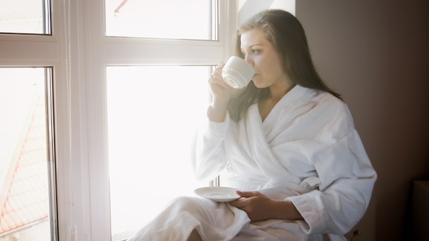 Piękna Młoda Kobieta Siedzi W Pokoju Hotelowym Przy Dużym Oknie I Pije Kawę