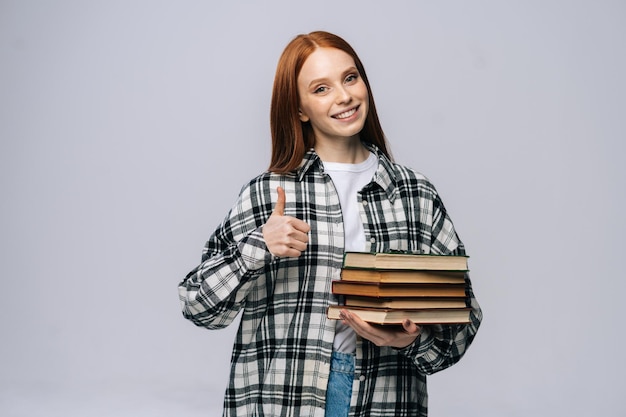 Piękna młoda kobieta studentka gospodarstwa książki i pokazując kciuk do góry gest patrząc na kamery na szarym tle na białym tle. Ładna rudowłosa modelka emocjonalnie pokazująca mimikę.