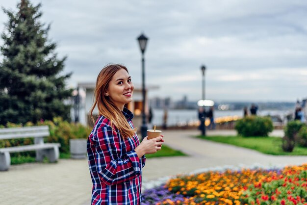 Piękna młoda kobieta trzyma filiżankę kawy i uśmiecha się w parku