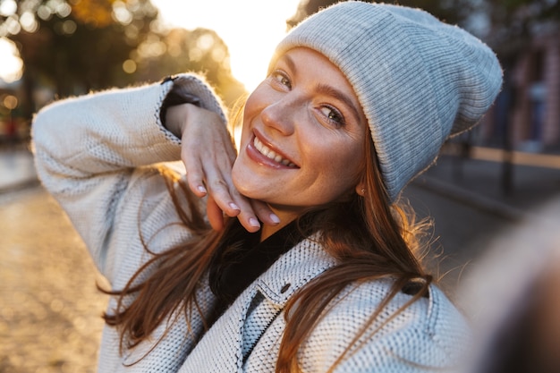 Piękna młoda kobieta ubrana w jesienny płaszcz i kapelusz spaceru na świeżym powietrzu, biorąc selfie