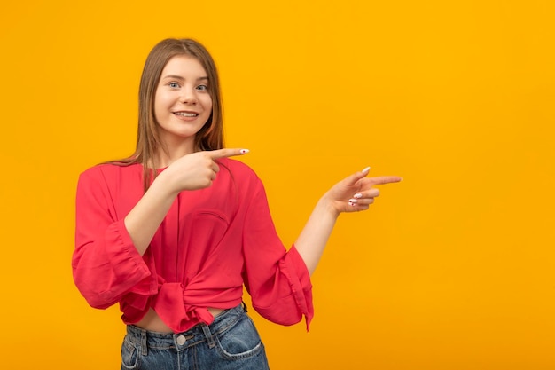 Piękna młoda kobieta uśmiecha się i wskazuje palcem na bok Izoluj na pomarańczowym tle Patrzy w kamerę Skopiuj miejsce