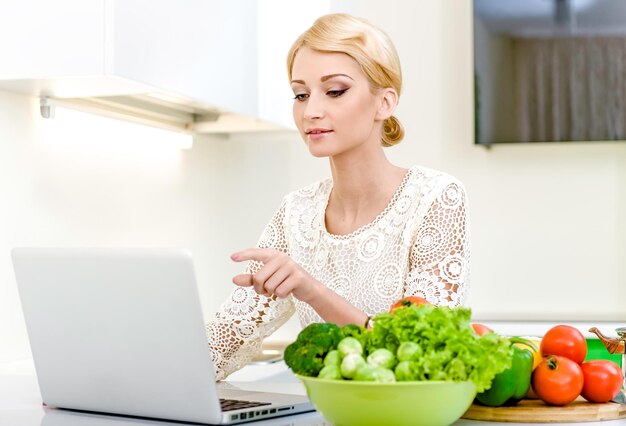Piękna młoda kobieta używająca laptopa, siedząc w domu z świeżymi warzywami