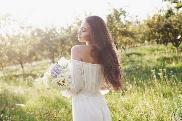 Piękna młoda kobieta w białej lekkiej sukience i bukiet letnich kwiatów spędza piękny dzień w ogrodzie