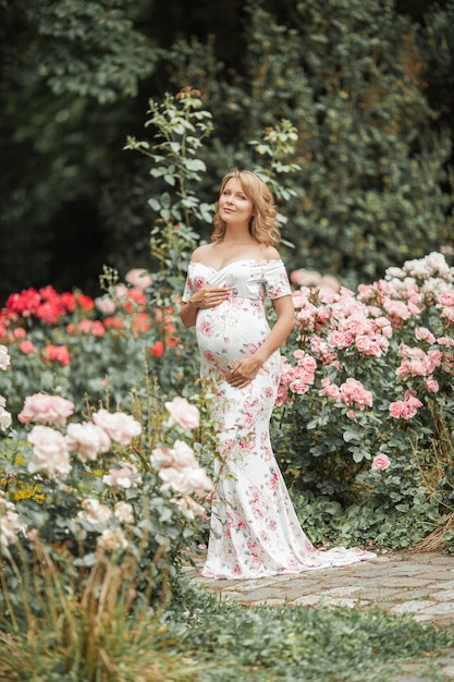 Piękna młoda kobieta w ciąży idzie w ogrodzie różanym. Portret kobiety w ciąży w sukience. Lato.