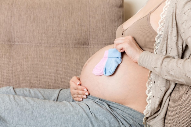 Piękna młoda kobieta w ciąży trzyma parę ślicznych skarpetek dziecięcych na brzuchu, leżąc na kanapie w domu