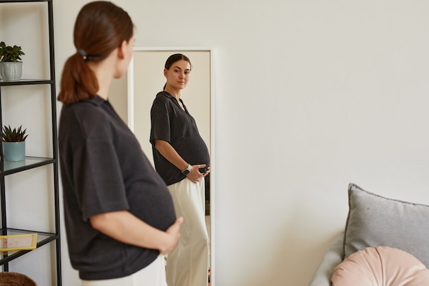 Zdjęcie piękna młoda kobieta w ciąży w wygodnym noszeniu, stojąca w przytulnym salonie i patrząca na brzuch w lustrze