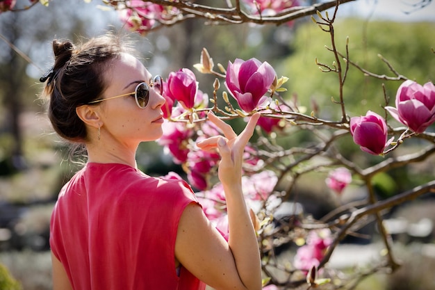 Piękna młoda kobieta w czerwonej sukience i okularach przeciwsłonecznych podziwia czerwoną kwitnącą magnolię w wiosennym ogrodzie