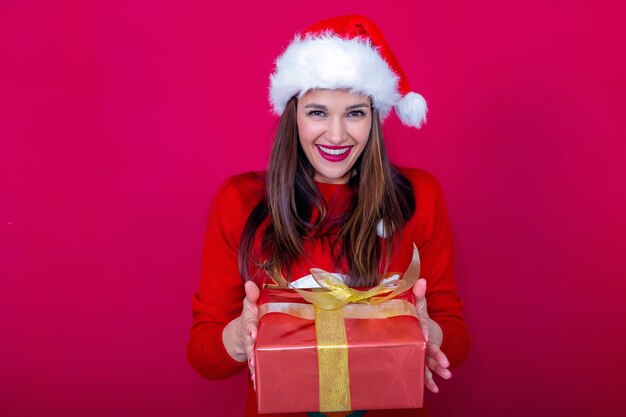 Piękna młoda kobieta w czerwonym swetrze i czapce Świętego Mikołaja oferująca prezent z szerokim uśmiechem