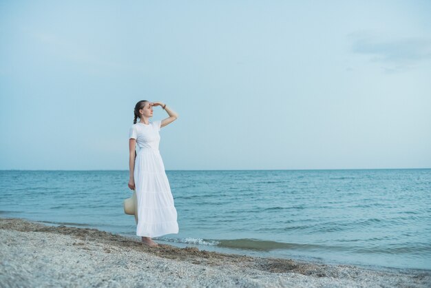 Piękna młoda kobieta w długiej białej sukni stoi na plaży i patrzy w dal.