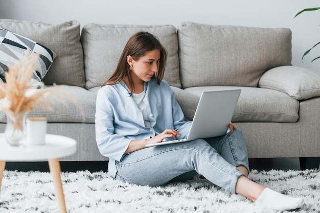 Piękna młoda kobieta w dżinsach i niebieskiej koszuli siedzi na podłodze z laptopem w domu w domu