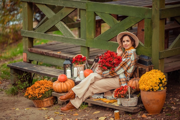 Piękna młoda kobieta w kapeluszu na spacerze jesienią w lesie z kwiatami i dyniami na tle drewnianej altany