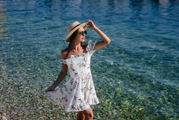 Piękna młoda kobieta w kapeluszu, okularach i lekkiej sukience przechadza się brzegiem oceanu na tle ogromnych skał w słoneczny dzień. Turystyka i wyjazdy turystyczne.