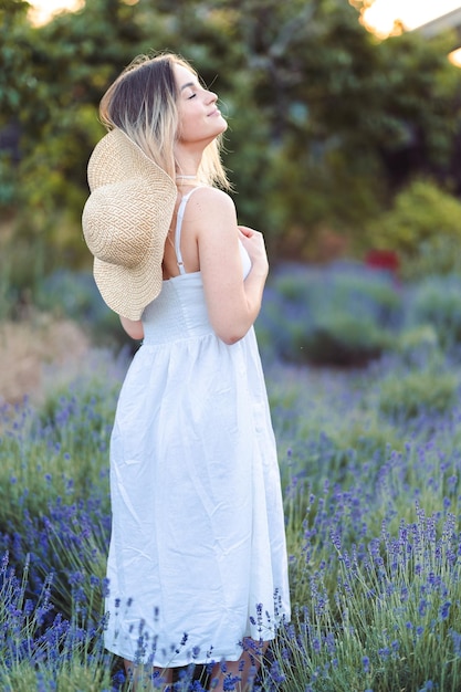Piękna młoda kobieta w letniej sukience i słomkowym kapeluszu wdycha świeży zapach lawendy