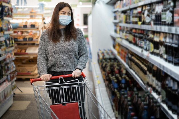 Piękna młoda kobieta w masce ochronnej idzie do supermarketu w pobliżu półek z metalowym wózkiem na zakupy Zakupy w dużym sklepie