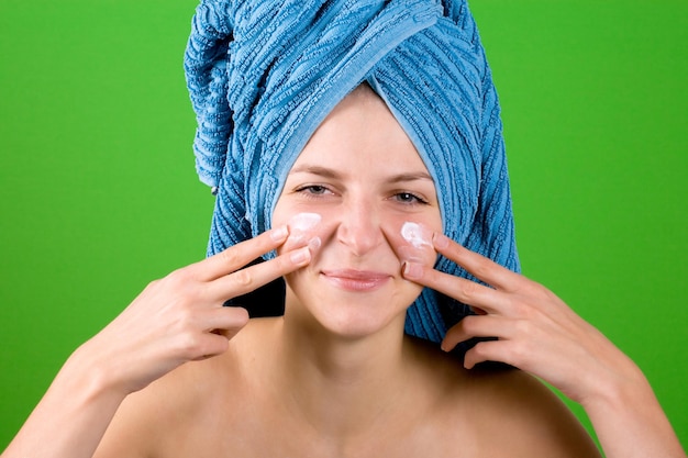Zdjęcie piękna młoda kobieta w niebieskim ręczniku nakładająca krem do twarzy na zielonym tle