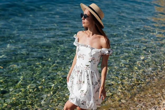 Piękna młoda kobieta w okularach kapeluszowych i lekkiej sukience spaceruje brzegiem oceanu na tle ogromnych skał w słoneczny dzień Turystyka i wyjazdy turystyczne