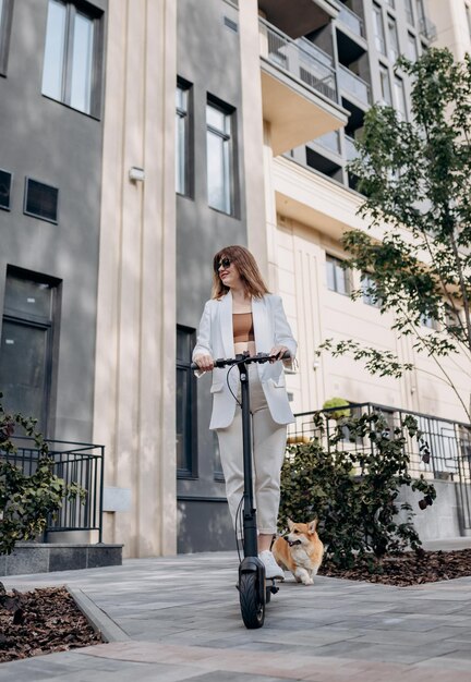 Piękna młoda kobieta w okularach przeciwsłonecznych i białym garniturze jedzie na swoim elektrycznym skuterze z psem Corgi w pobliżu nowoczesnego budynku w mieście i odwraca wzrok