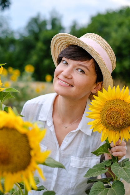 Piękna młoda kobieta w słomkowym kapeluszu stoi latem na polu słoneczników i słodko się uśmiecha.