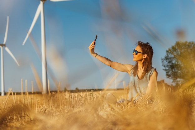 Zdjęcie piękna młoda kobieta w średnim wieku na polu zbóż robi selfie z wiatrakami w tle i okularami przeciwsłonecznymi