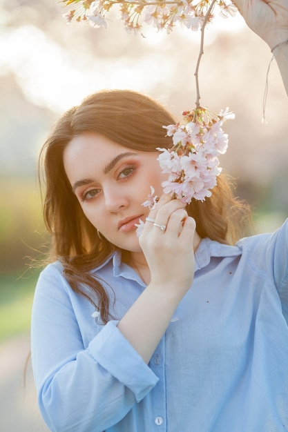 Piękna młoda kobieta w wieku 19 lat w lekkich, casualowych ubraniach w pastelowych odcieniach w pobliżu kwitnących drzew sakura Wiosenny portret modelki o niebieskich oczach