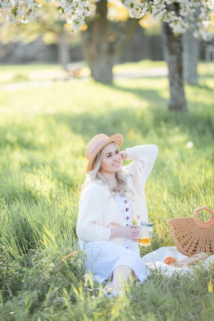 Piękna młoda kobieta w wiklinowym kapeluszu odpoczywa na pikniku w kwitnącym ogrodzie. Białe kwiaty. Wiosna. Szczęście.