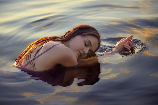 Piękna młoda kobieta w wodzie jeziora w letniej sukience o zachodzie słońca Portret romantycznej mokrej dziewczyny o zachodzie słońca ciepłe słońce naturalne piękno kobiety