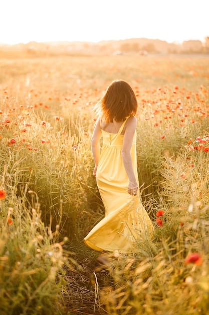 Piękna młoda kobieta w żółtej sukience spaceru w polu maku w letni dzień. Dziewczyna z kwiatami na wsi. Selektywne skupienie