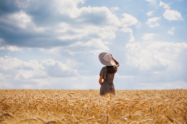 Piękna młoda kobieta z brązowym słuchem w różanej sukience i kapeluszu, ciesząca się świeżym powietrzem, patrząca na horyzont na idealnym polu pszenicy