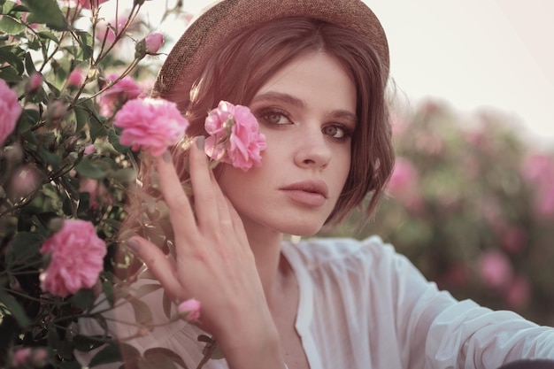 Piękna młoda kobieta z kręconymi włosami pozuje w pobliżu róż w ogrodzie Koncepcja reklamy perfum