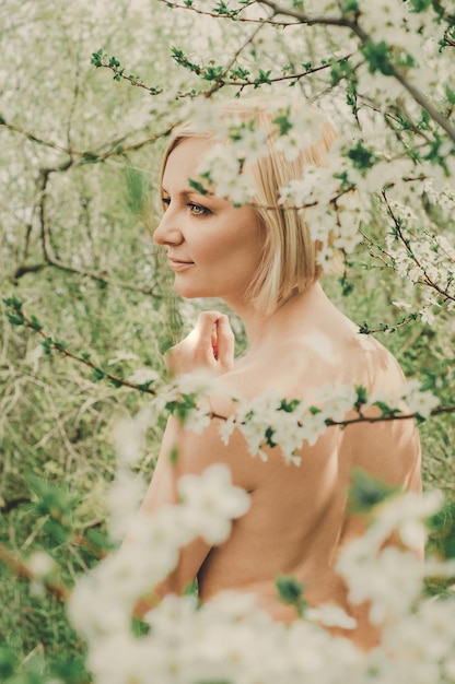 Piękna młoda kobieta z krótką blond fryzurą, nagimi ramionami i plecami spogląda w dal wśród kwitnących wiśni w słonecznym ogrodzie wiosną.