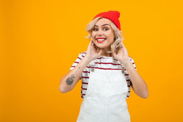 Piękna młoda kobieta z krótkimi blond kręconymi włosami i jasnym makijażem w białym kombinezonie i czerwonym kapeluszu gestykuluje i uśmiecha się, portret na pomarańczowo