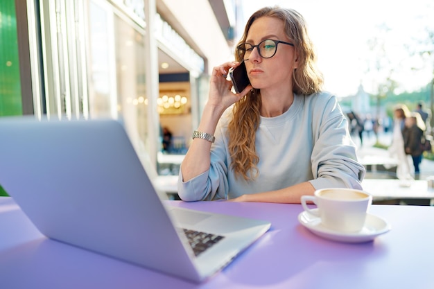 Piękna młoda kobieta z laptopem rozmawia przez telefon w kawiarni na świeżym powietrzu