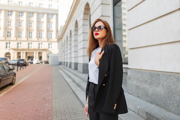 Piękna młoda kobieta z modnymi okularami przeciwsłonecznymi w modnych modnych ubraniach z czarnym blazerem chodzi po mieście w pobliżu starego budynku