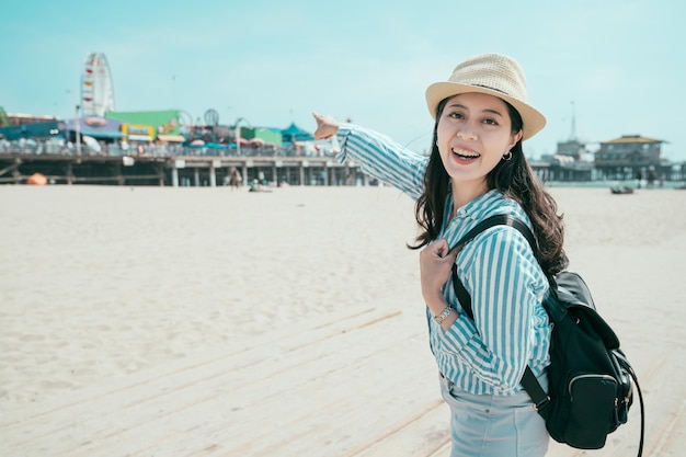 Piękna młoda kobieta z plecakiem zabawy w parku rozrywki. relaks na wiosnę koncepcja piaszczystej plaży. ładna dziewczyna podróżnik w słomkowym kapeluszu patrząca na twarz aparat uśmiechający się punktowy park rozrywki w słoneczny dzień