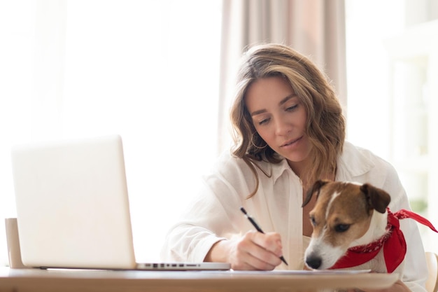 Piękna młoda kobieta z uroczym psem pracującym na laptopie w domu