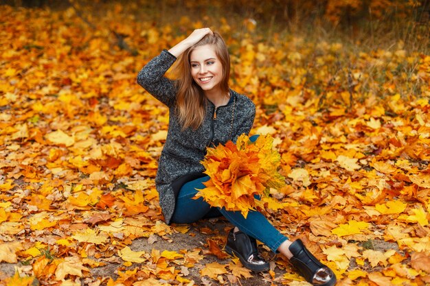 Piękna młoda kobieta z uśmiechem w stylowym płaszczu siedzi na żółtych liściach jesienią